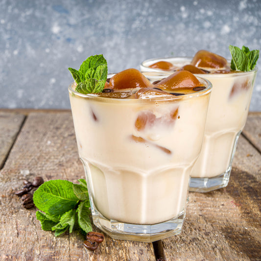Two glasses of iced coffee with cream and ice cubes sit on a wooden surface, garnished with fresh mint leaves and surrounded by coffee beans, against a textured gray background.