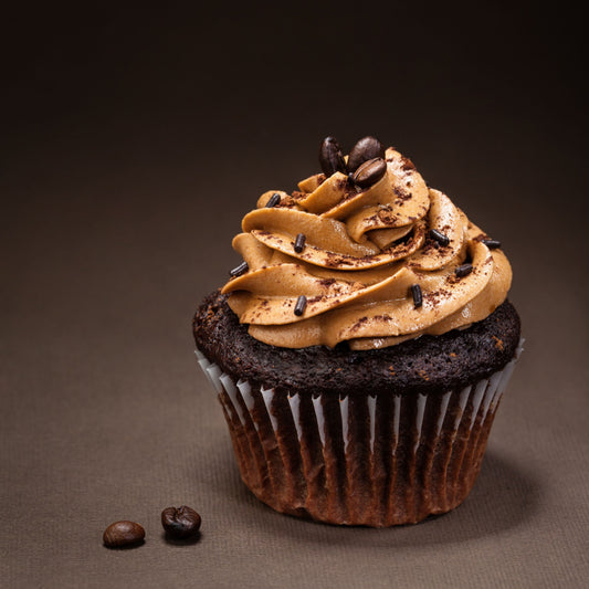 A chocolate cupcake topped with creamy coffee-flavored frosting, sprinkled with chocolate shavings and coffee beans, sits on a dark background.