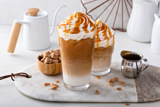 Two tall glasses of caramel iced coffee topped with whipped cream and drizzled with caramel sit on a marble serving board, accompanied by a bowl of sugar cubes and a small milk jug.