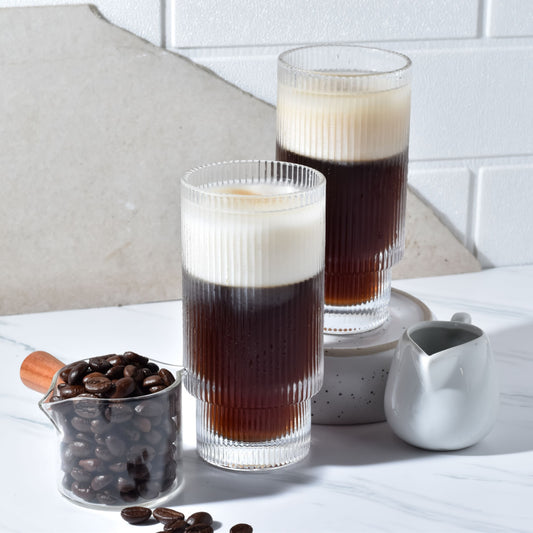 Two tall, ribbed glasses filled with dark coffee topped with frothy cream sit on a marble surface, accompanied by a small white creamer and a measuring cup of coffee beans.