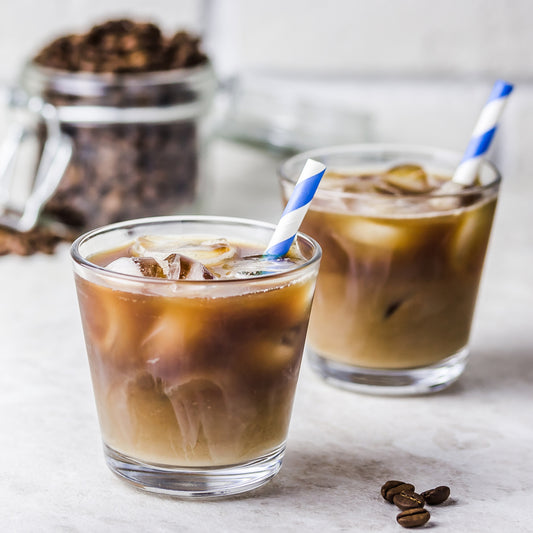 Two glasses of iced coffee with blue and white striped straws sit on a light surface, with a jar of coffee beans in the background and a few coffee beans scattered nearby.