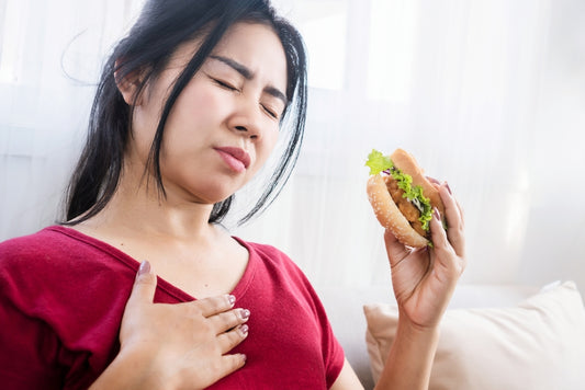 A woman in a red shirt holds a hamburger while pressing her chest with a pained expression, sitting on a couch in a bright room with soft curtains.