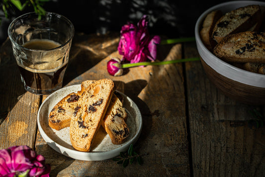 A small plate with slices of biscotti sits on a wooden table, next to a glass of dark coffee and pink flowers, with sunlight casting soft shadows across the scene.