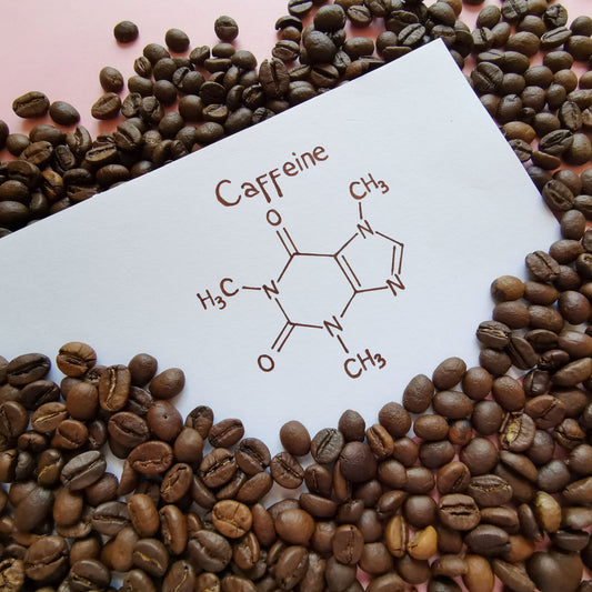 A white card displaying the molecular structure of caffeine is surrounded by scattered coffee beans on a pink background.