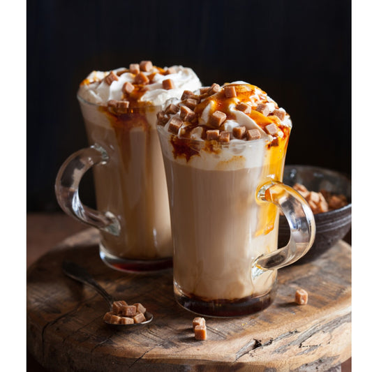 Two clear glass mugs filled with creamy coffee topped with whipped cream, caramel drizzle, and caramel bits, sit on a wooden surface alongside a spoon and a small bowl of caramel pieces.