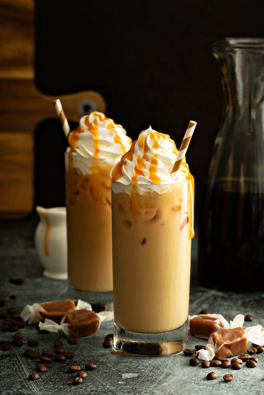 Two tall glasses of iced coffee topped with whipped cream and caramel drizzle sit on a dark surface, surrounded by coffee beans and caramel candies, with a glass pitcher in the background.