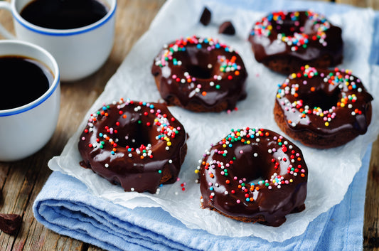 Five chocolate-glazed donuts with colorful sprinkles are arranged on parchment paper, accompanied by two cups of coffee on a wooden table, creating a cozy snack setting.