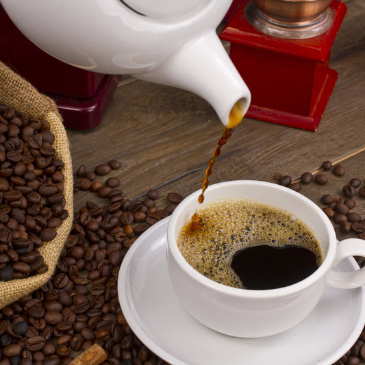 A white teapot pours hot coffee into a white cup sitting on a saucer, surrounded by scattered coffee beans and a burlap sack, with a red coffee grinder in the background.