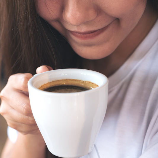 A woman smiles softly while holding a white cup of coffee close to her face, enjoying the aroma in a cozy indoor setting.