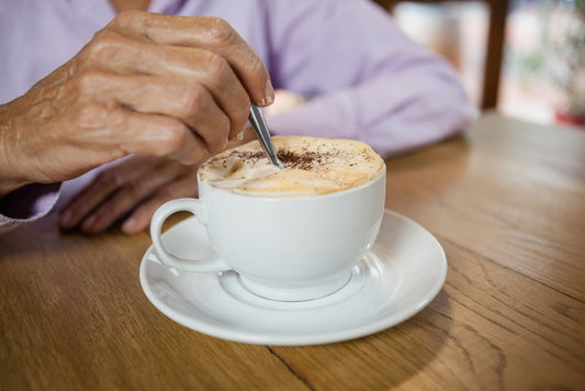 A hand stirs a frothy cappuccino in a white cup on a wooden table, with a soft purple shirt visible in the background, suggesting a cozy café setting.