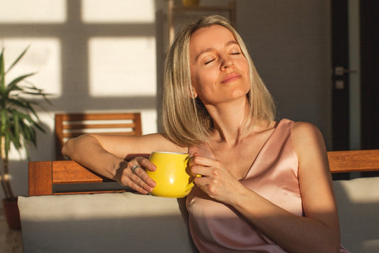 A woman with shoulder-length blonde hair sits on a couch, holding a yellow mug and smiling softly, enjoying sunlight streaming through a window in a cozy, modern living space.