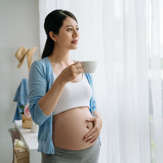 A pregnant woman in a light blue cardigan stands by a window, holding a cup, with a thoughtful expression as she gazes outside, surrounded by soft natural light.