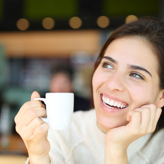 A young woman smiles brightly while holding a small white coffee cup, resting her chin on her hand, in a cozy café setting with soft lighting in the background.