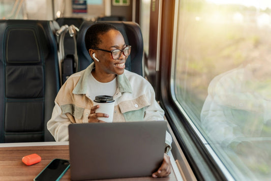 A smiling person with glasses and earbuds sits in a train seat, holding a coffee cup while working on a laptop, with sunlight streaming through the window.