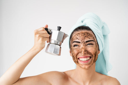 A woman with a towel on her head smiles widely while holding a moka pot in one hand, her face covered in coffee grounds, against a plain white background.