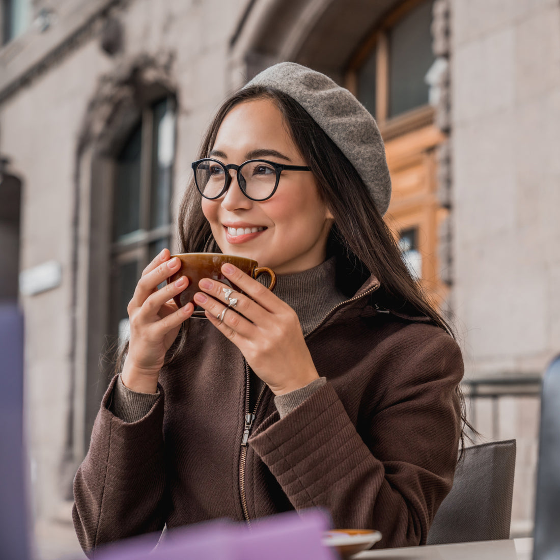 A woman with long hair and glasses smiles while holding a brown cup, sitting at an outdoor café with a stone building in the background, wearing a brown jacket and gray beret.