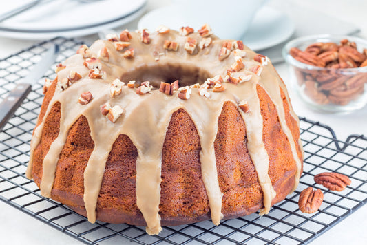 A freshly baked bundt cake with a glossy caramel glaze and chopped pecans sits on a cooling rack, with a bowl of whole pecans and plates in the background.