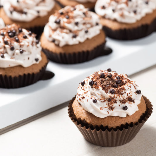 A close-up of a chocolate cupcake topped with swirls of white frosting, chocolate shavings, and dark sprinkles, placed among other similar cupcakes in a white box.