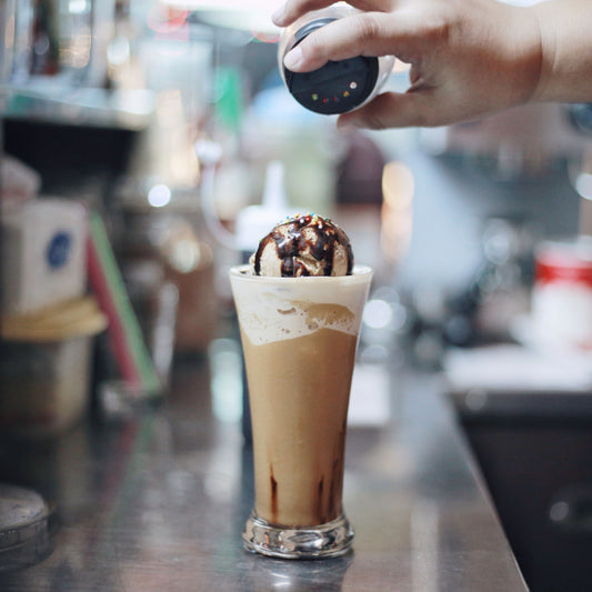 A hand sprinkles colorful toppings over a tall glass filled with a frothy coffee drink topped with a scoop of ice cream and drizzled with chocolate sauce, set against a blurred café background.