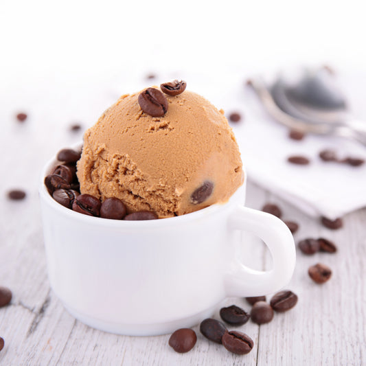 A scoop of coffee-flavored ice cream sits in a white cup, surrounded by scattered coffee beans on a light wooden surface, with a spoon and napkin blurred in the background.