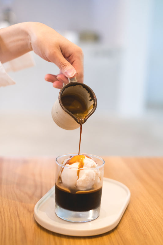 A hand pours a rich caramel sauce from a small pitcher over a glass of vanilla ice cream sitting on a light wooden table, with a soft-focus background.