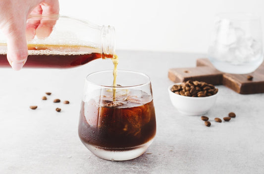 A hand pours cold brew coffee from a glass bottle into a round glass filled with ice, with coffee beans scattered on a light gray surface and a small bowl of beans nearby.
