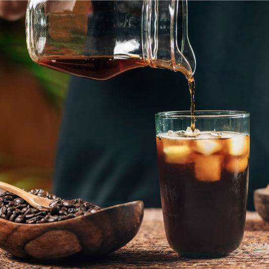 A person pours cold brew coffee from a glass pitcher into a tall glass filled with ice, while a wooden bowl of coffee beans sits nearby on a textured surface.