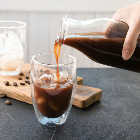 A hand pours cold brew coffee from a glass bottle into a clear glass filled with ice, with coffee beans scattered on a dark surface and another glass in the background.