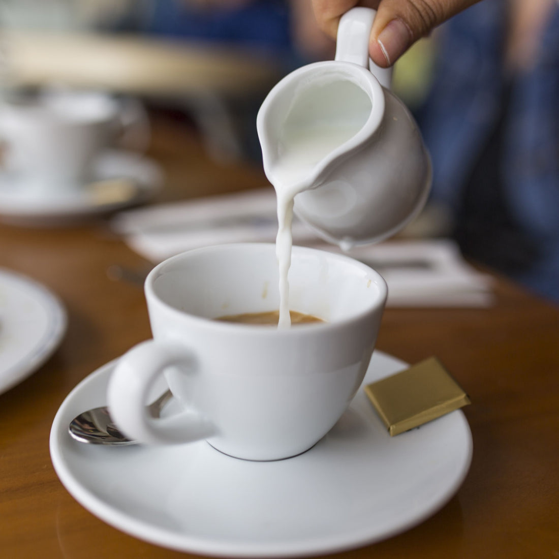 A hand pours milk from a small white creamer into a white coffee cup on a saucer, with a gold chocolate square nearby, set on a wooden table in a casual dining environment.