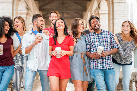 A diverse group of eight young adults joyfully laugh and walk together, holding coffee cups, under an arched walkway in a sunny outdoor setting.