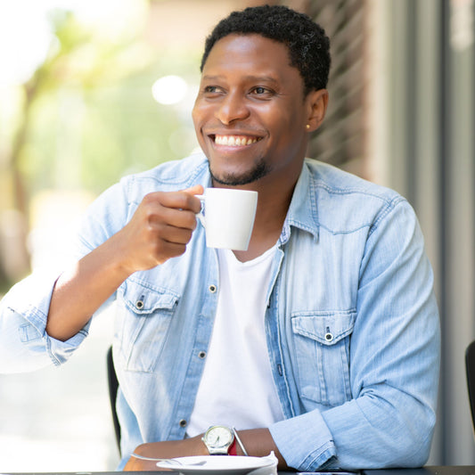 A smiling man holds a white coffee cup while sitting at a table outdoors, wearing a denim shirt over a white t-shirt, with sunlight and greenery visible in the background.