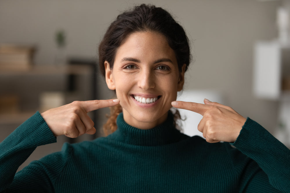 A woman in a green sweater smiles brightly while pointing to her own cheeks, set against a softly blurred indoor background.