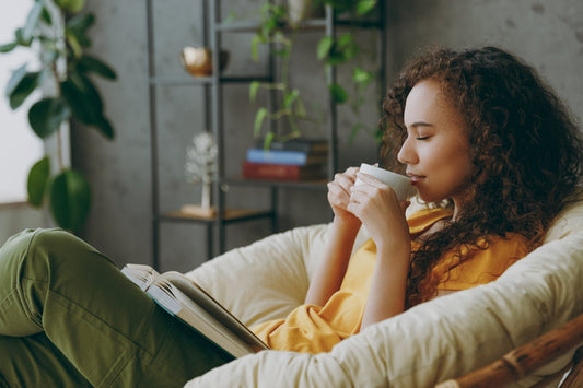 A woman with curly hair sits comfortably in a cozy chair, sipping from a cup while reading a book in a bright room filled with plants and shelves.