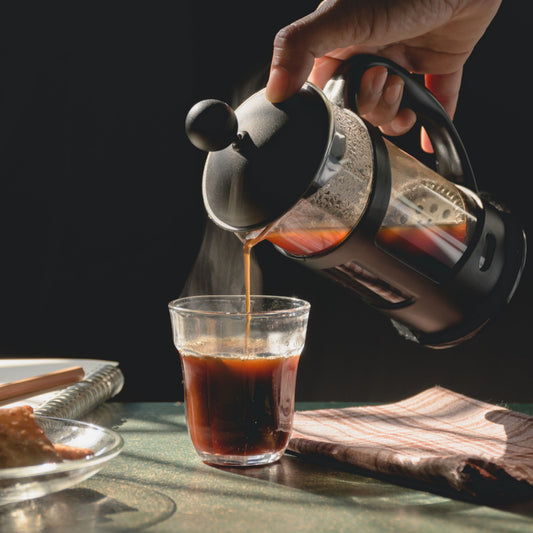 A hand pours steaming coffee from a French press into a clear glass on a table, with a patterned cloth and a plate of snacks nearby, set against a dark background.