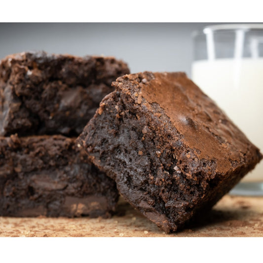 Three rich, fudgy brownies are stacked on a wooden surface, with a glass of milk in the background, creating a tempting dessert scene.