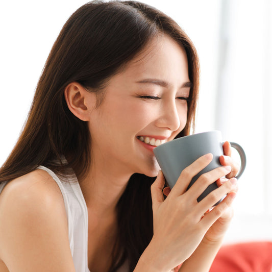 A woman with long hair smiles joyfully while holding a gray mug close to her face, enjoying a moment of warmth and comfort in a bright, cozy indoor setting.