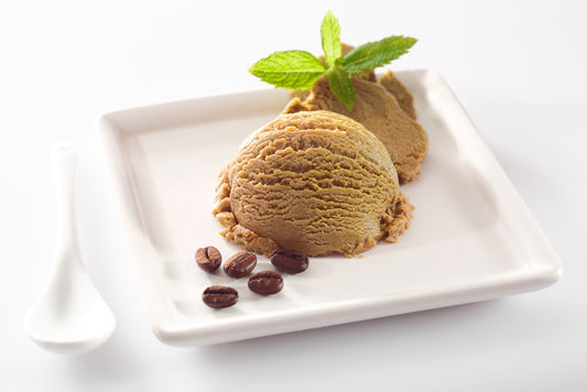 A scoop of green tea ice cream is served on a white plate, garnished with a mint leaf, alongside a few coffee beans and a white spoon.