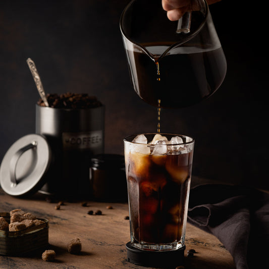 A hand pours dark coffee from a glass pitcher into a tall glass filled with ice, surrounded by coffee beans, a sugar container, and a dark cloth on a wooden table.