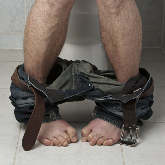 A person sits on a toilet with their jeans and belt around their ankles, exposing their bare feet and legs in a bathroom setting.