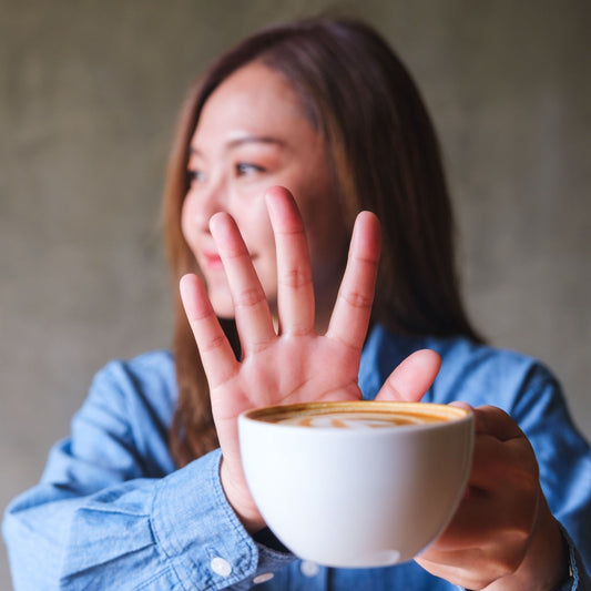 A woman in a blue shirt holds a white coffee cup while playfully raising her hand in a stop gesture, with a blurred gray background.