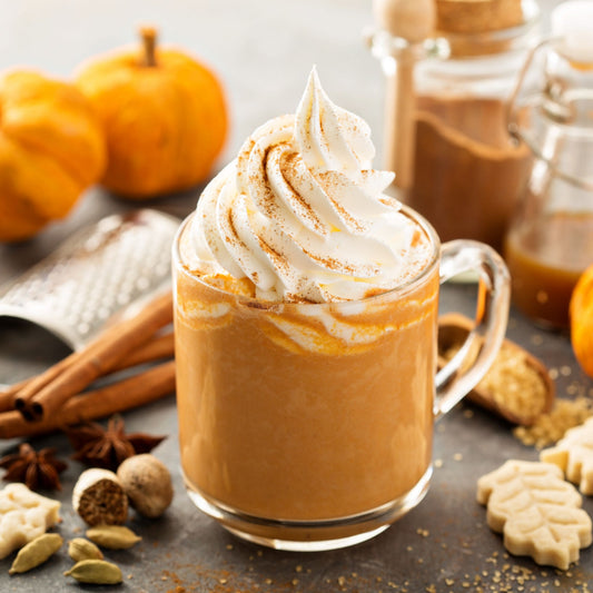 A clear glass mug filled with creamy pumpkin spice latte topped with whipped cream and cinnamon sits on a table surrounded by small pumpkins, spices, and autumn-themed cookies.