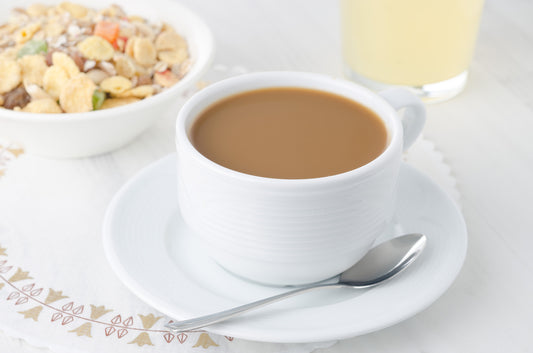 A white cup of coffee sits on a saucer with a spoon beside it, next to a bowl of cereal containing various ingredients, all arranged on a light-colored table.