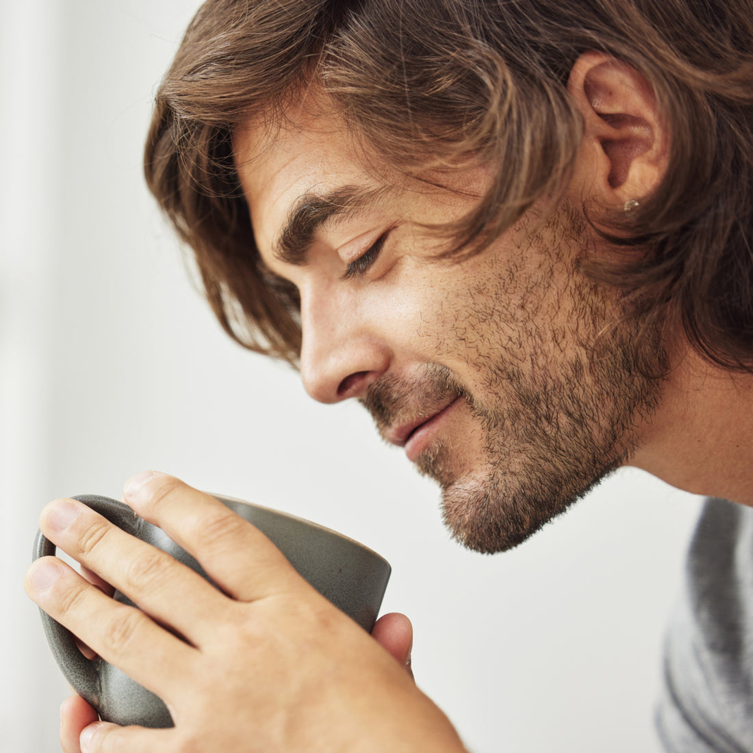 A man with tousled hair and a light beard gently holds a gray mug close to his face, smiling softly as he enjoys a warm beverage in a bright, minimalistic setting.
