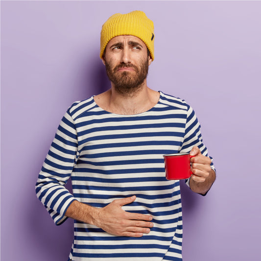 A man with a beard and a yellow beanie looks concerned while holding a red mug and resting his hand on his stomach against a light purple background.