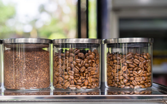 Three glass jars with metal lids are filled with different coffee products: ground coffee on the left, roasted coffee beans in the middle, and another type of roasted beans on the right, set against a blurred outdoor background.