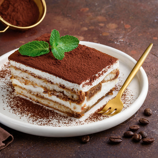 A slice of tiramisu topped with cocoa powder and a mint leaf is served on a white plate, accompanied by a gold fork, with coffee beans scattered on a rustic brown surface.