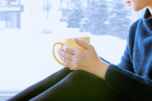 A person sits by a snowy window, holding a yellow mug with both hands, wearing a cozy blue sweater, and appears to be enjoying a quiet moment indoors.
