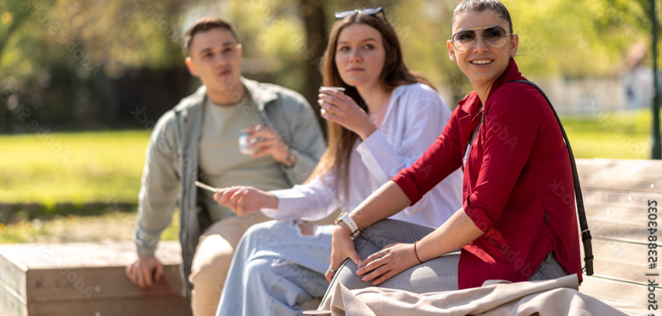 Three young adults sit on a bench outdoors in a park, enjoying drinks on a sunny day. The woman in front wears sunglasses and a red shirt, while the others chat in the background. Trees and greenery are visible.