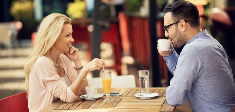 A woman and a man sit at an outdoor café table, smiling and talking. The woman stirs her drink while the man holds a coffee cup. There are glasses of water, orange juice, and a smartphone on the table.