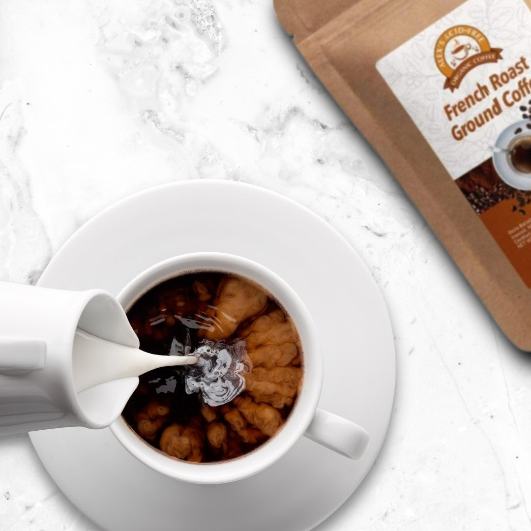 Cream swirls into a white cup of Alex's Low-Acid Organic Coffee™ - French Roast Fresh Ground (5lbs) as it sits on a saucer, with the brown bag from Alex's Low Acid Coffee nearby on a white marble countertop.